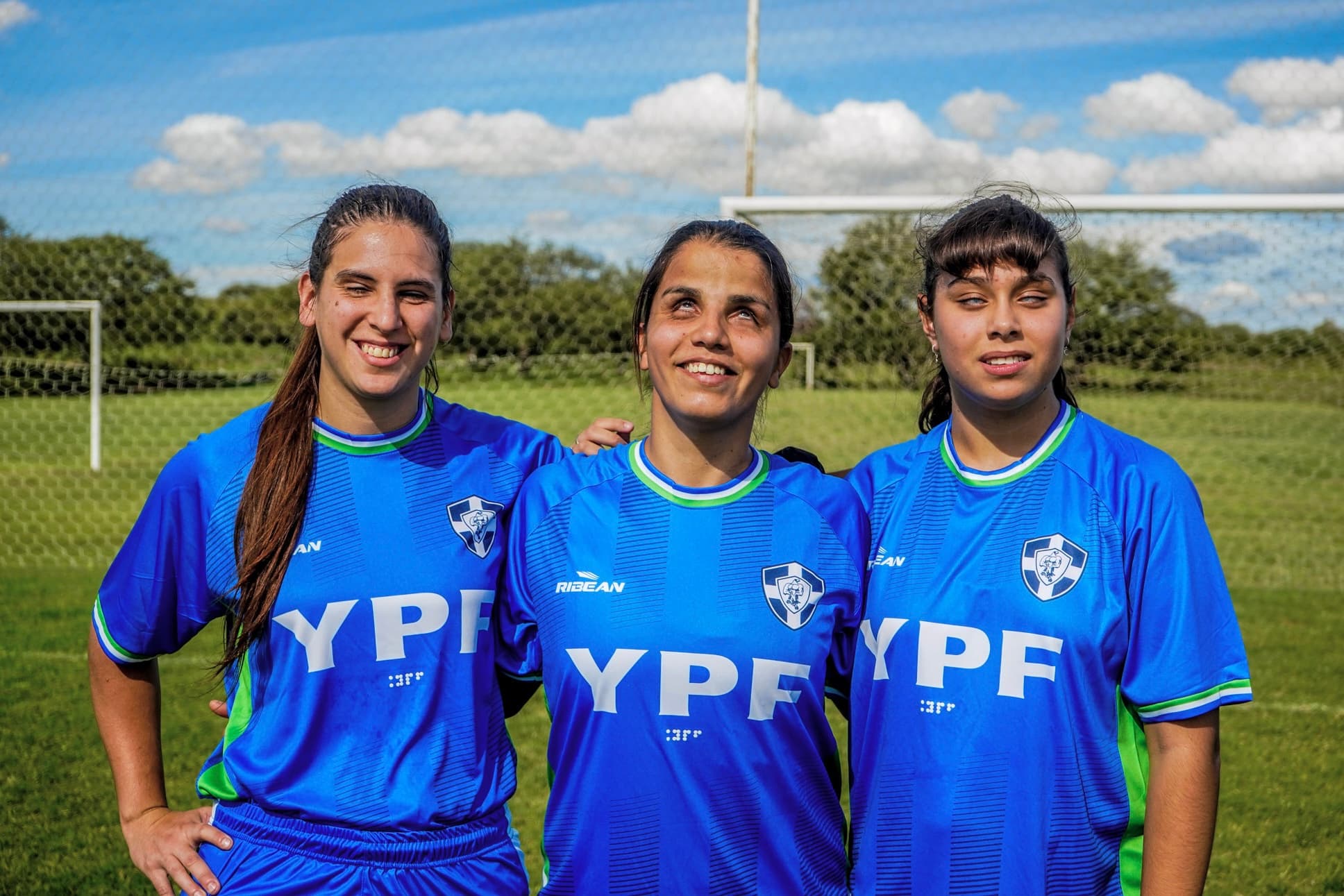 Tres jugadoras de fútbol para ciegos abrazadas y sonriendo en la cancha, con la camiseta azul del club.