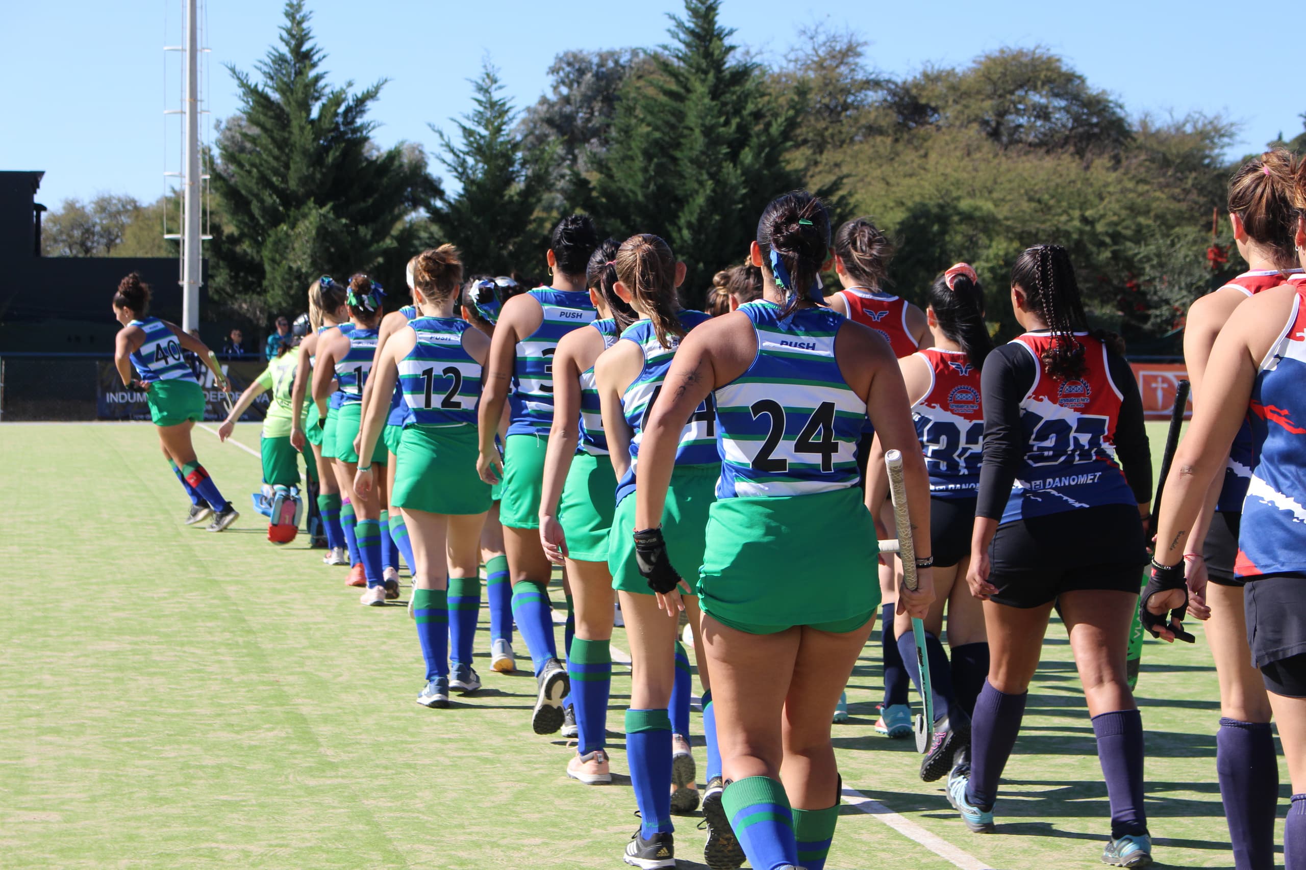 El plantel de hockey entra a la cancha en fila, de espaldas, con la camiseta a rayas verdes y azules del club.