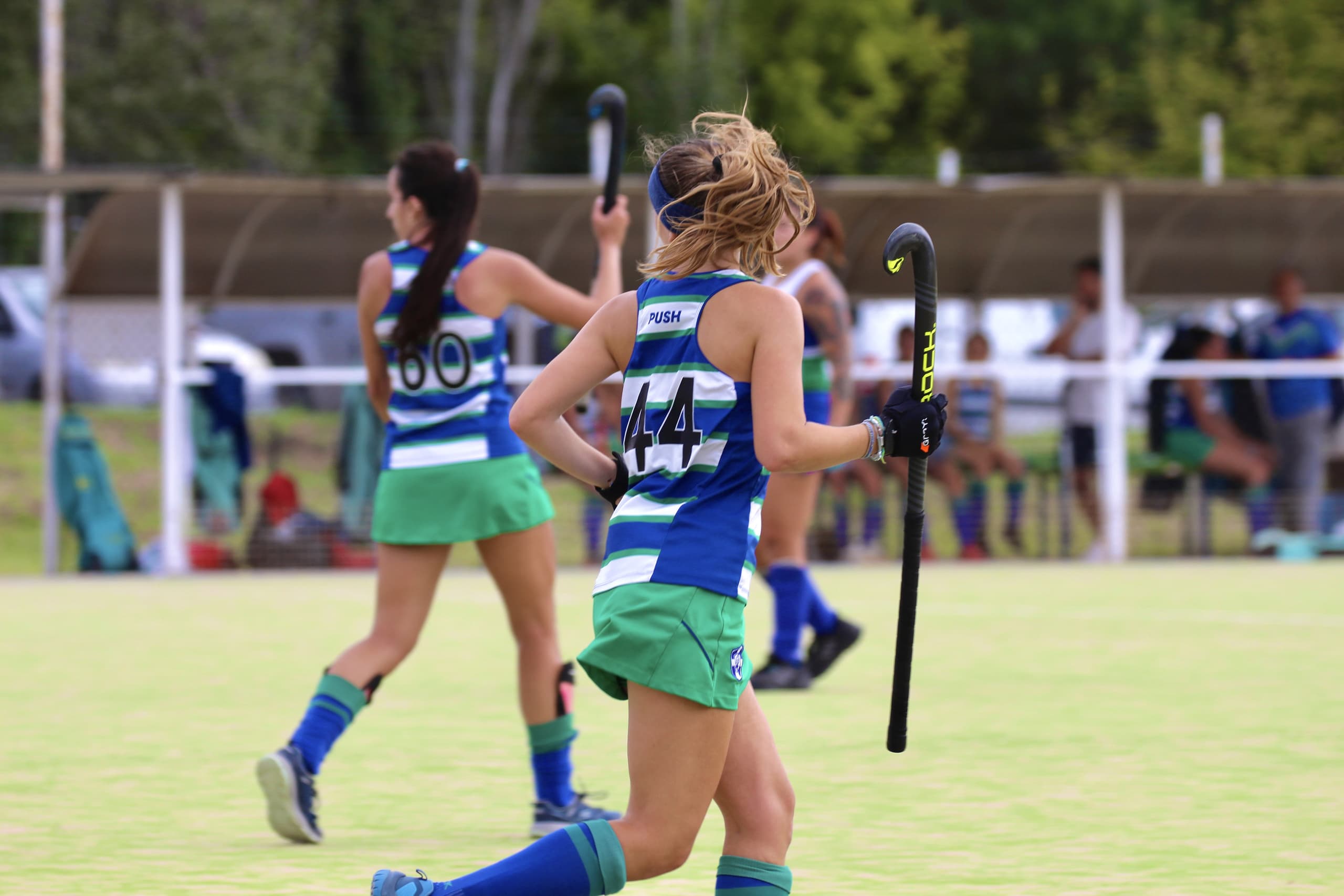 Dos jugadoras corren por la cancha durante un partido de hockey, con el palo en alto.
