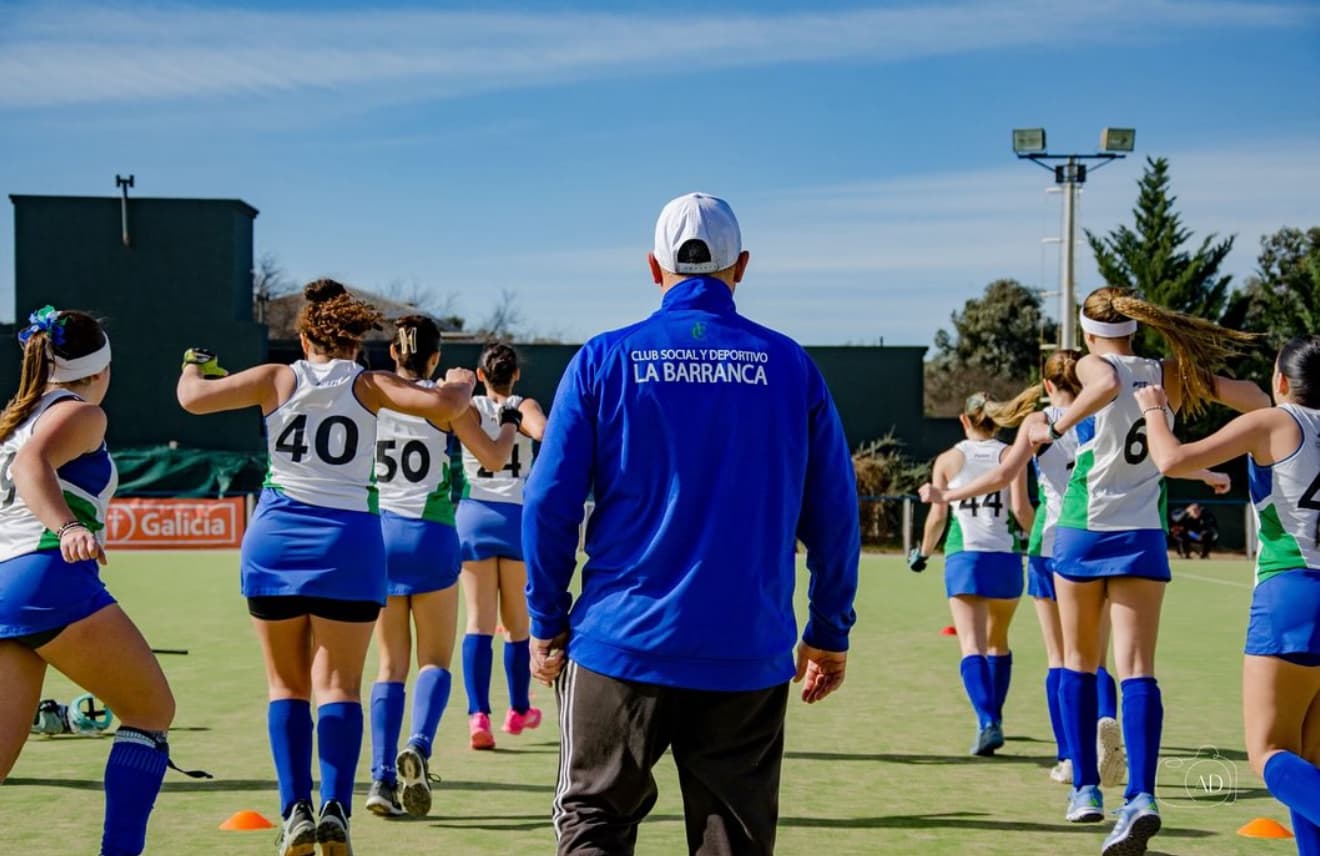 El entrenador, de espaldas y con la campera del club, dirige la entrada en calor del plantel de hockey.