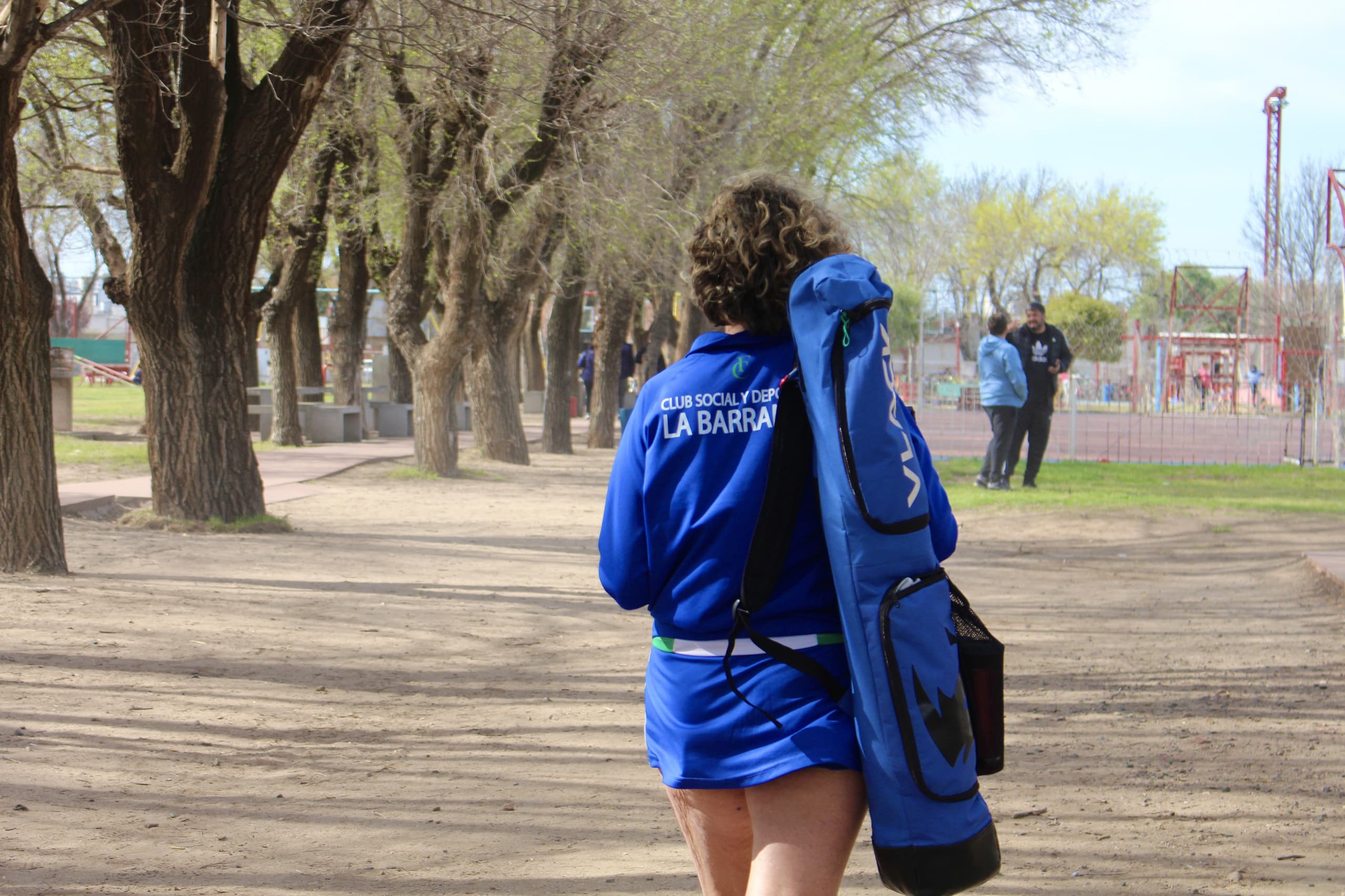 Una jugadora camina de espaldas por la arboleda del predio con el bolso de palos al hombro y la campera del club.